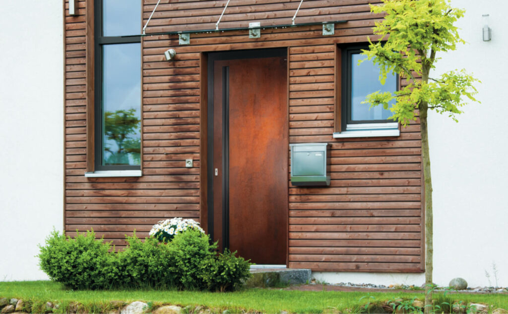 Moderne Holz-Haustür in brauner Fassade mit Glasvordach, seitlichem Fenster und Vorgarten mit Natursteinmauer und Baum